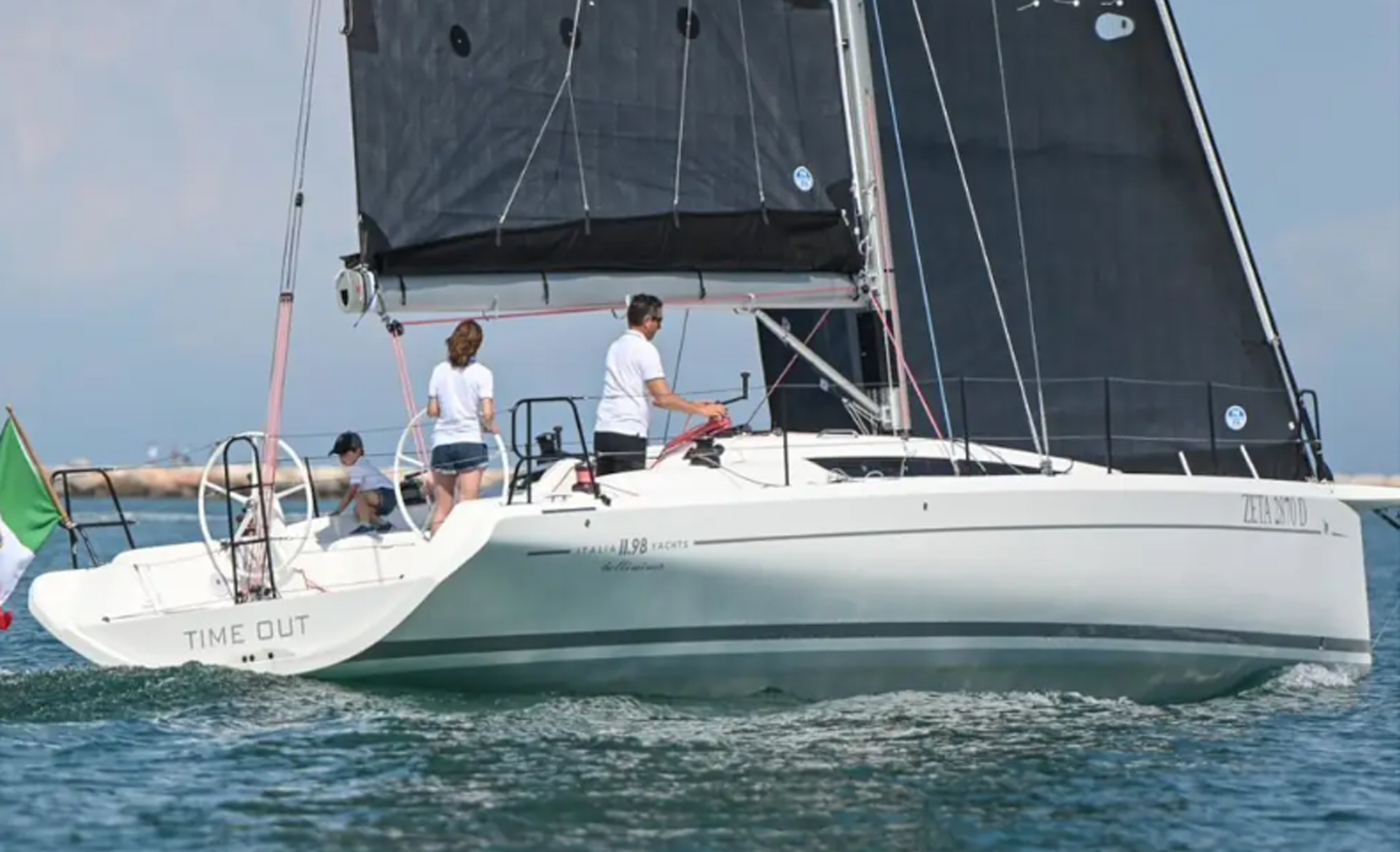 Side view of the Italia Yachts IY 11.98 Bellissima sailing yacht with crew onboard and a black mainsail.