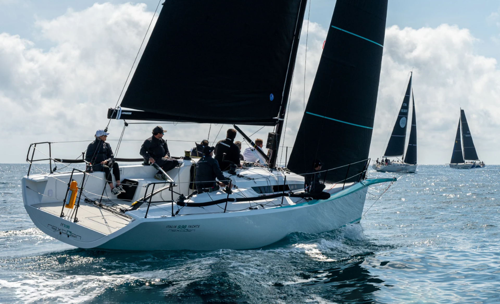 Italia Yachts IY 9.98 nextGen sailing yacht underway with dark racing sails and crew members on deck.